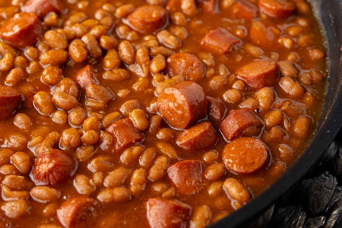 Klassisches Rezept für Würstchen mit Bohnen 2 Eine Nahaufnahme von gebackenen Bohnen, gemischt mit geschnittenen Würstchen in einer braunen Sauce, in einer Pfanne.