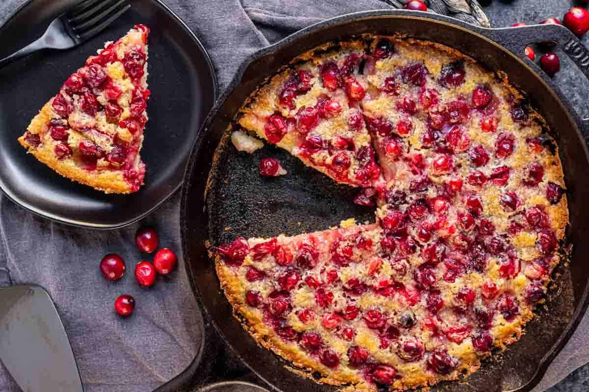 Cranberry-Streuselkuchen 2 Eine Kirsch-bedeckte Nachspeise in einer Pfanne, mit einem Stück, das entfernt und auf einen schwarzen Teller daneben gelegt wurde, umgeben von verstreuten frischen Kirschen.