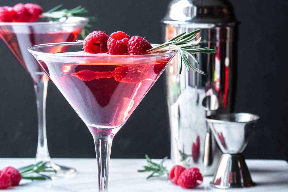 **Ein Galentine’s-Schmaus für deine Mädels** 2 Zwei pinkfarbene Cocktails mit Himbeeren und Rosmarin als Garnitur in Martinigläsern; ein Cocktailshaker und Messbecher im Hintergrund.