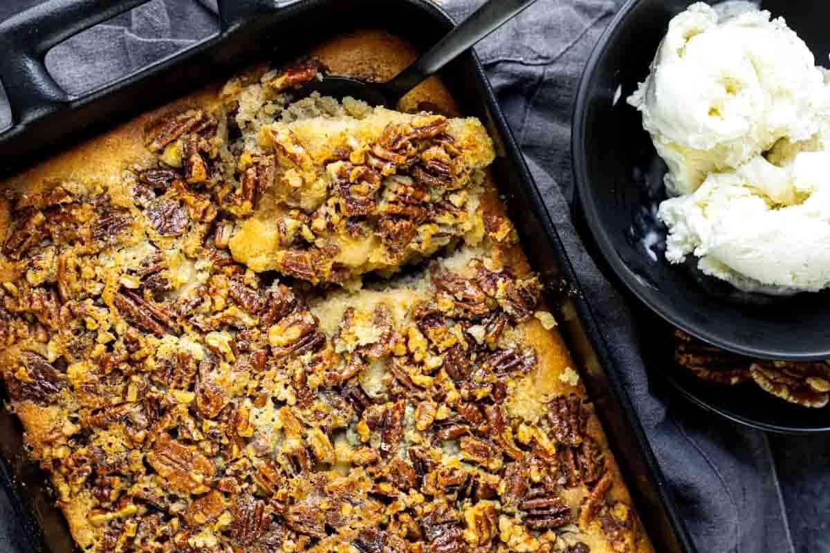 Unwiderstehliche Desserts mit Pekannusskuchenfüllung 2 Eine Auflaufform mit Pekannuss-Cobbler und einem Servierlöffel, daneben eine schwarze Schale mit Vanilleeis.