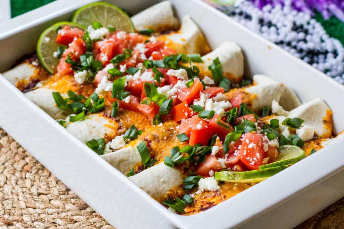 Festliche Abendessenrezepte für den Cinco de Mayo 2 Ein Blech Enchiladas, belegt mit Käse, gewürfelten Tomaten, Frühlingszwiebeln und Limettenspalten, serviert auf einem geflochtenen Platzset.