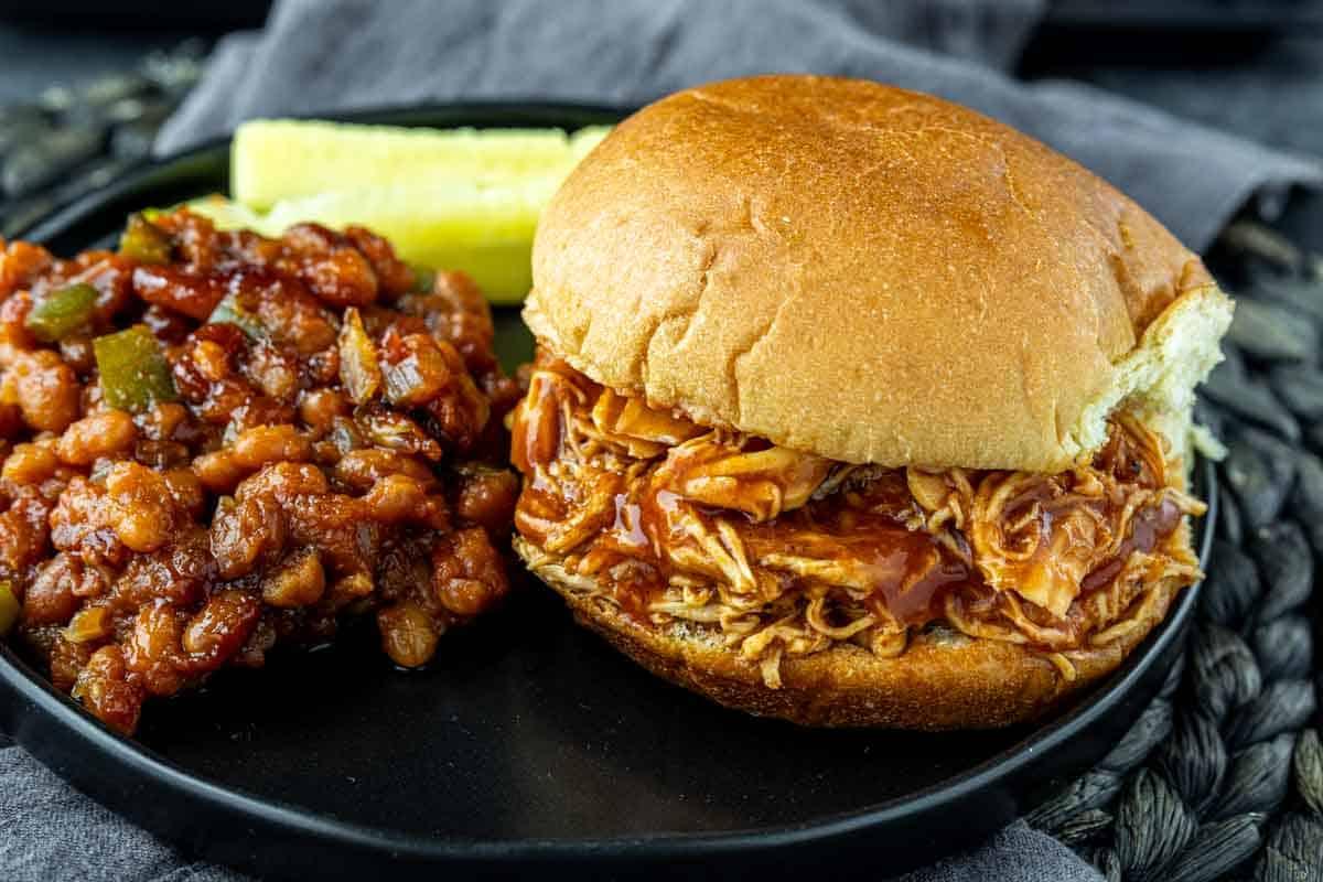 BBQ Pulled Chicken im Slow Cooker 2 Ein gerupftes Barbecue-Hähnchen-Sandwich liegt auf einem schwarzen Teller neben einer Portion gebackener Bohnen und einer Gewürzgurkenscheibe.