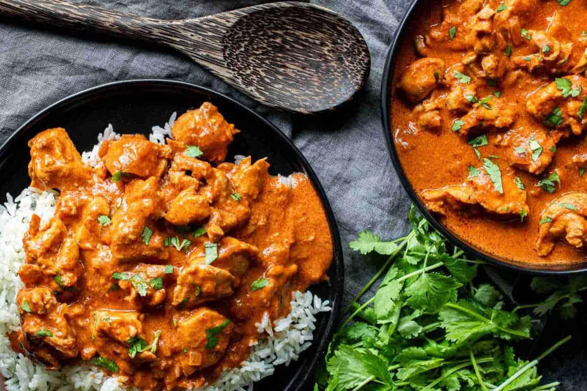 **Schnelle und einfache Hähnchengerichte für vielbeschäftigte Familien** 2 Ein Teller Hähnchen Tikka Masala, serviert über Reis, garniert mit Kräutern, daneben ein Holzlöffel und frischer Koriander auf einer dunklen, strukturierten Oberfläche.