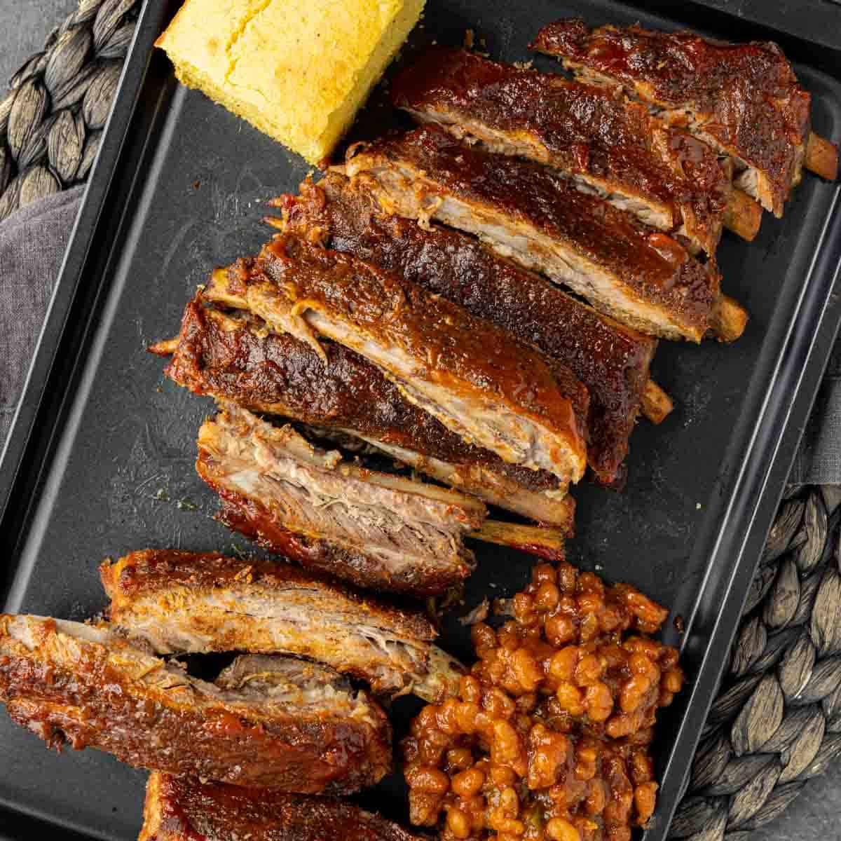 St. Louis Style Spareribs: Langsam gegarte Perfektion 2 Geschnittene St. Louis-Style Ribs auf einem schwarzen Teller mit Maisbrot und gebackenen Bohnen, von oben gesehen.