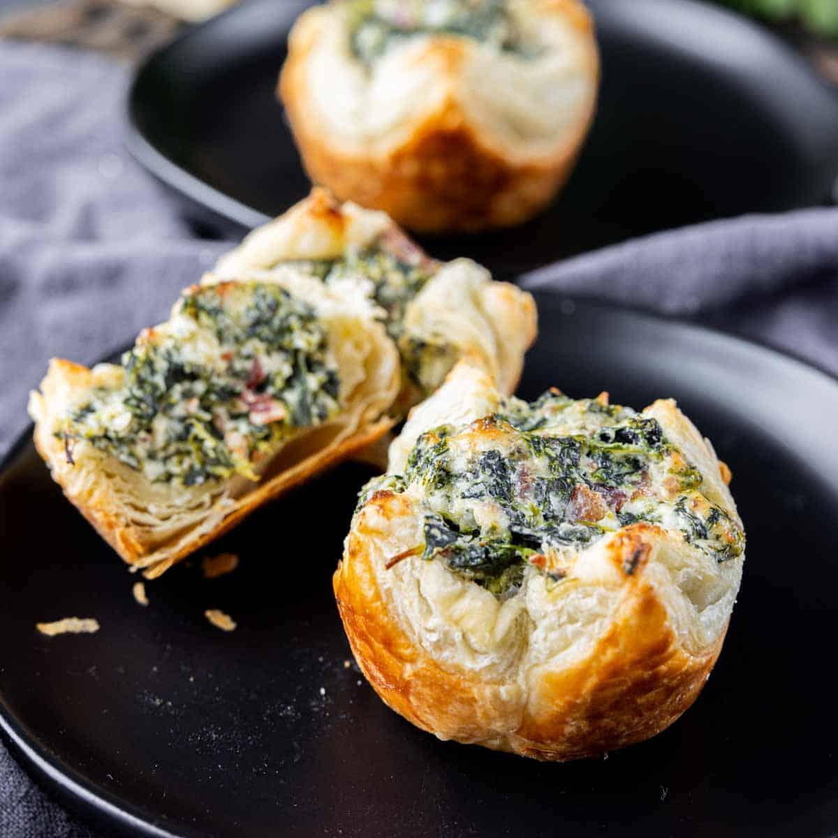 Diese einfachen Spinat-Blätterteig-Häppchen sind eines meiner Lieblings-Fingerfoods, die man herzhaft vorbereiten kann. Diese mundgerechten Becher sind mit Rahmspinat und Speck gefüllt. Sie eignen sich perfekt zum Servieren an Thanksgiving, Weihnachten oder Silvesterpartys. #partyessen #spinat #vorspeisen #mundgerecht #speck #weihnachten #silvester #blätterteig #hausgemachtesinteresse Spinat-Blätterteig-Häppchen auf einem schwarzen Teller.