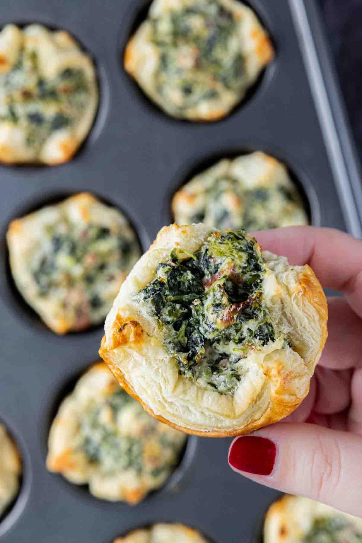 Spinat-Blätterteig-Vorspeise 11 Eine Hand hält ein Spinat-Blätterteig-Häppchen in einem Muffinblech.