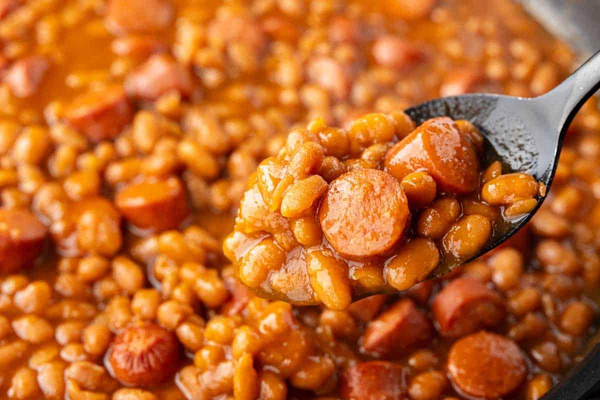 33 einfache und günstige Gerichte (die Ihre Familie auch wirklich essen möchte) 12 Nahaufnahme eines Löffels gebackener Bohnen und geschnittener Würstchen in einer dicken Soße, mit mehr Bohnen und Würstchen im Hintergrund.