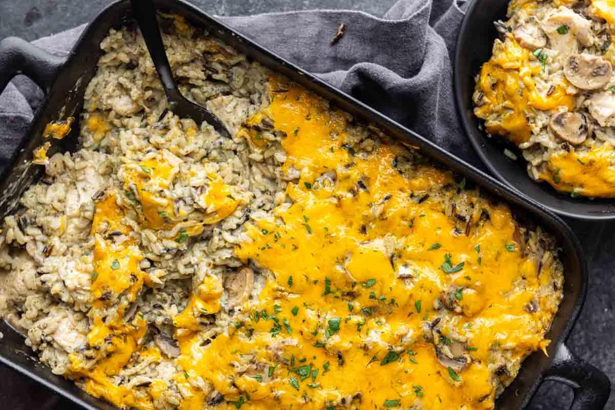 33 einfache und günstige Gerichte (die Ihre Familie auch wirklich essen möchte) 14 Eine Auflaufform gefüllt mit käsigem, gebackenem Pilz- und Reisauflauf, wobei eine Portion entnommen wurde und ein Servierlöffel in der Pfanne liegt.