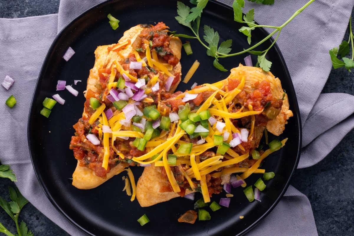 **Schnelle und einfache Hähnchengerichte für vielbeschäftigte Familien** 14 Zwei gefüllte Tacos, belegt mit Käse, gewürfelten Zwiebeln und geschnittenen grünen Chilischoten, serviert auf einem schwarzen Teller mit frischem Koriander und einer grauen Serviette.