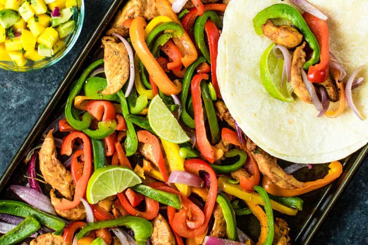Festliche Abendessenrezepte für den Cinco de Mayo 15 Bunte Hähnchen-Fajitas mit Paprika, Zwiebeln und Limettenscheiben auf einem Backblech, begleitet von einer Tortilla und einer Beilage Mango-Salsa.