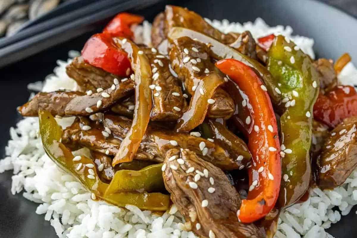 33 einfache und günstige Gerichte (die Ihre Familie auch wirklich essen möchte) 16 Ein Teller mit weißem Reis, belegt mit gebratenen Rinderstreifen, roter und grüner Paprika, Zwiebeln, brauner Soße und Sesamkörnern.
