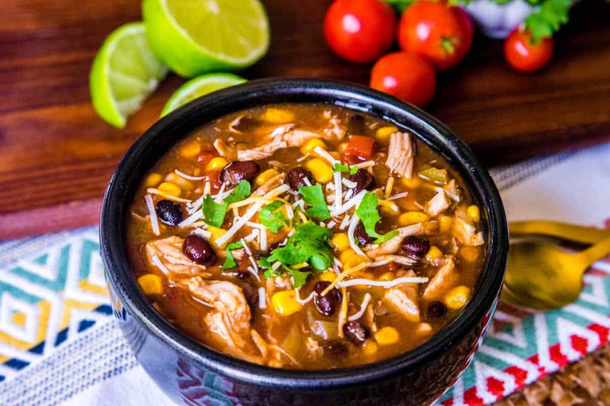 Festliche Abendessenrezepte für den Cinco de Mayo 16 Eine Schüssel Hühnchen-Tortilla-Suppe mit Bohnen und Mais, garniert mit geriebenem Käse und Koriander, begleitet von Limettenspalten und Kirschtomaten.