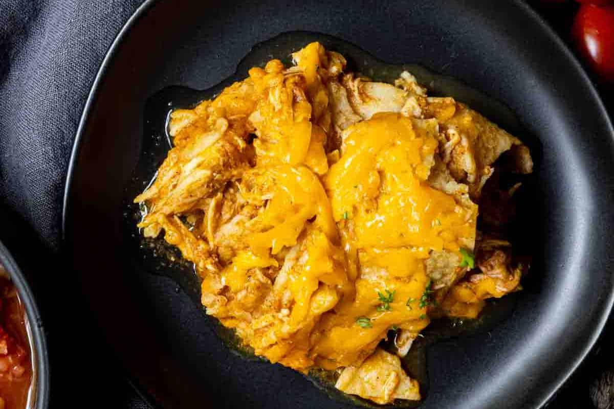 Festliche Abendessenrezepte für den Cinco de Mayo 17 Eine schwarze Pfanne mit zerkleinertem Hähnchen, belegt mit geschmolzenem Cheddar-Käse, garniert mit Kräutern, auf einem dunklen Hintergrund.