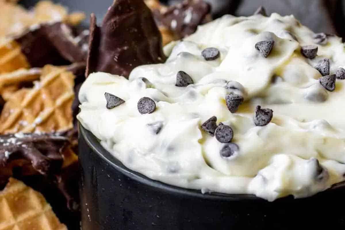 **Ein Galentine’s-Schmaus für deine Mädels** 20 Eine schwarze Schüssel, gefüllt mit cremigem weißen Schokoladenchip-Dip. Schokoladenüberzogene Waffelstücke sind im Hintergrund sichtbar.