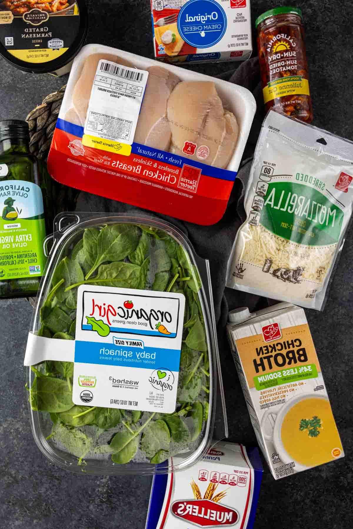 Cremige Florentiner Hähnchen-Pasta 3 Zutaten für Hähnchen Florentiner Auflauf, darunter Hähnchenbrust, Babyspinat, Mozzarella-Käse, Hühnerbrühe und andere Küchengrundnahrungsmittel, auf einer dunklen Oberfläche angeordnet.
