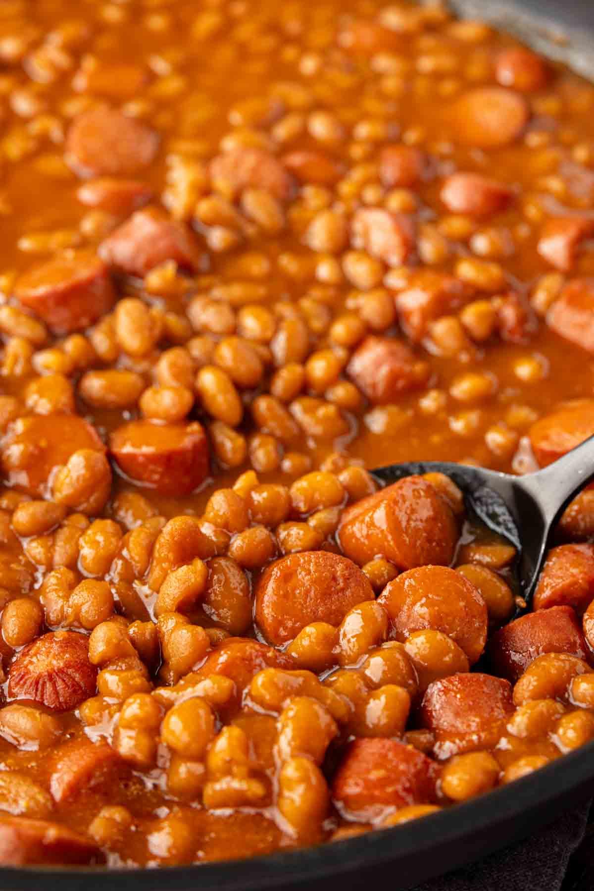 Klassisches Rezept für Würstchen mit Bohnen 3 Eine Pfanne gefüllt mit Würstchen und Bohnen. Ein Löffel liegt in der Mischung.