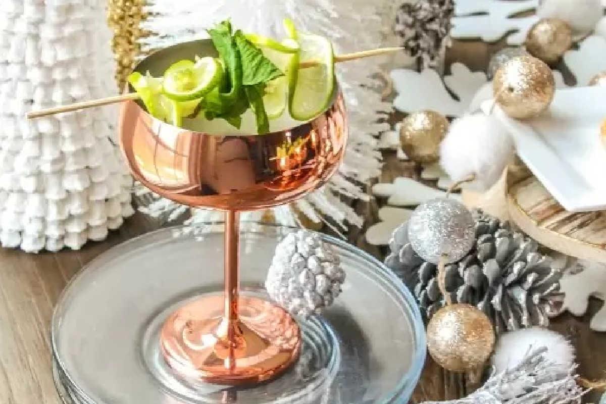 **Ein Galentine’s-Schmaus für deine Mädels** 3 Ein kupferfarbenes Cocktailglas, gefüllt mit Limette und Minze, auf einem Glasteller. Umgeben von Tannenzapfen und festlichen silbernen und weißen Dekorationen.
