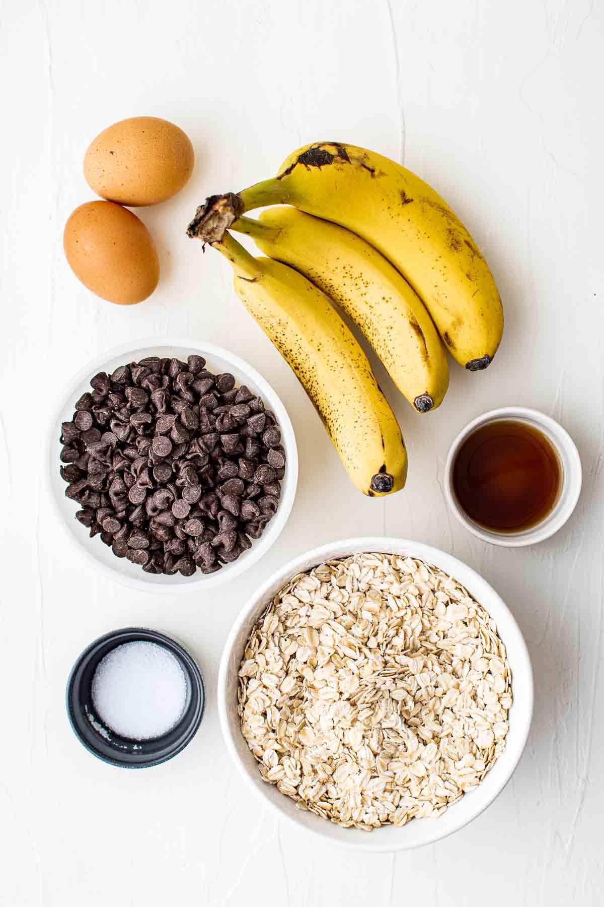 Gesunde Haferflockenkekse – Einfaches Frühstück oder Snack! 3 Zutaten auf einem Tisch: drei Bananen, zwei Eier, eine Schüssel Schokostückchen, eine Schüssel Haferflocken, eine kleine Schüssel Salz und eine kleine Schüssel Vanilleextrakt.