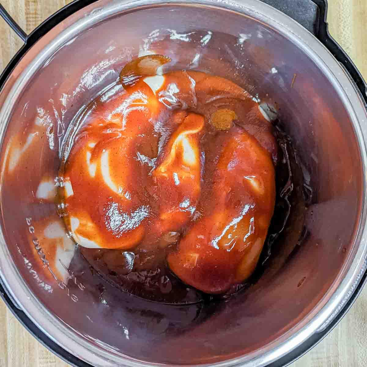 BBQ Pulled Chicken im Slow Cooker 3 Zwei rohe Hähnchenbrüste, bedeckt mit Barbecue-Soße, in einem Edelstahlschnellkochtopf.