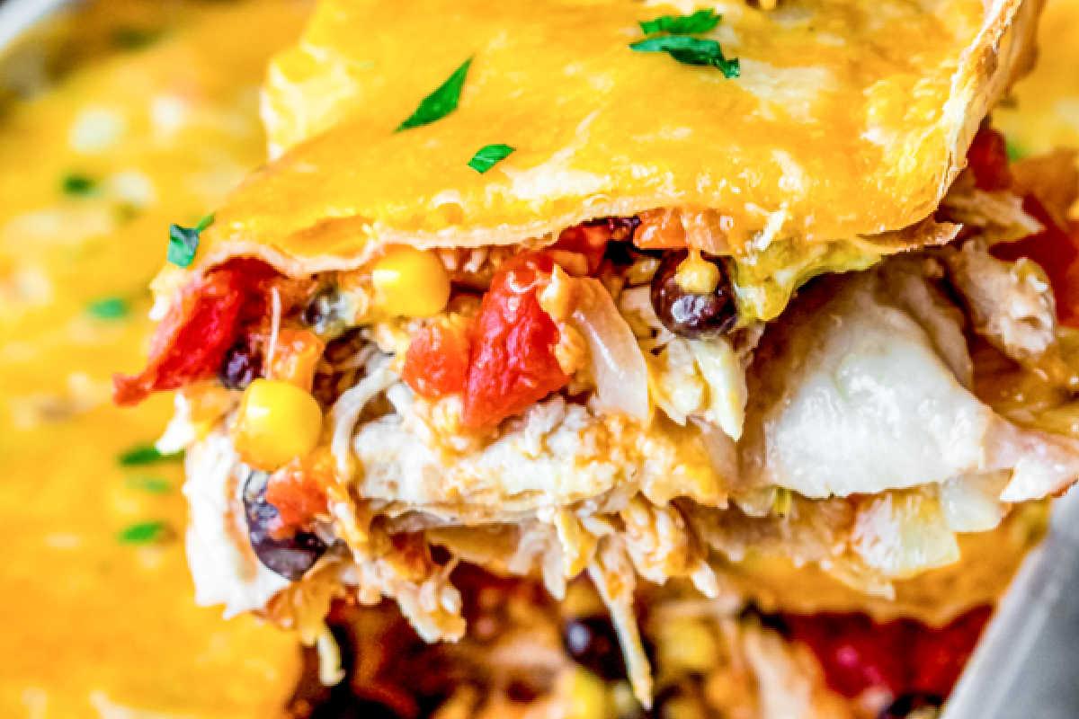 33 einfache und günstige Gerichte (die Ihre Familie auch wirklich essen möchte) 21 Nahaufnahme eines Stücks Hähnchen-Enchilada-Auflauf mit geschmolzenem Käse, gewürfelten Tomaten, schwarzen Bohnen, Mais und geschichteten Tortillas.