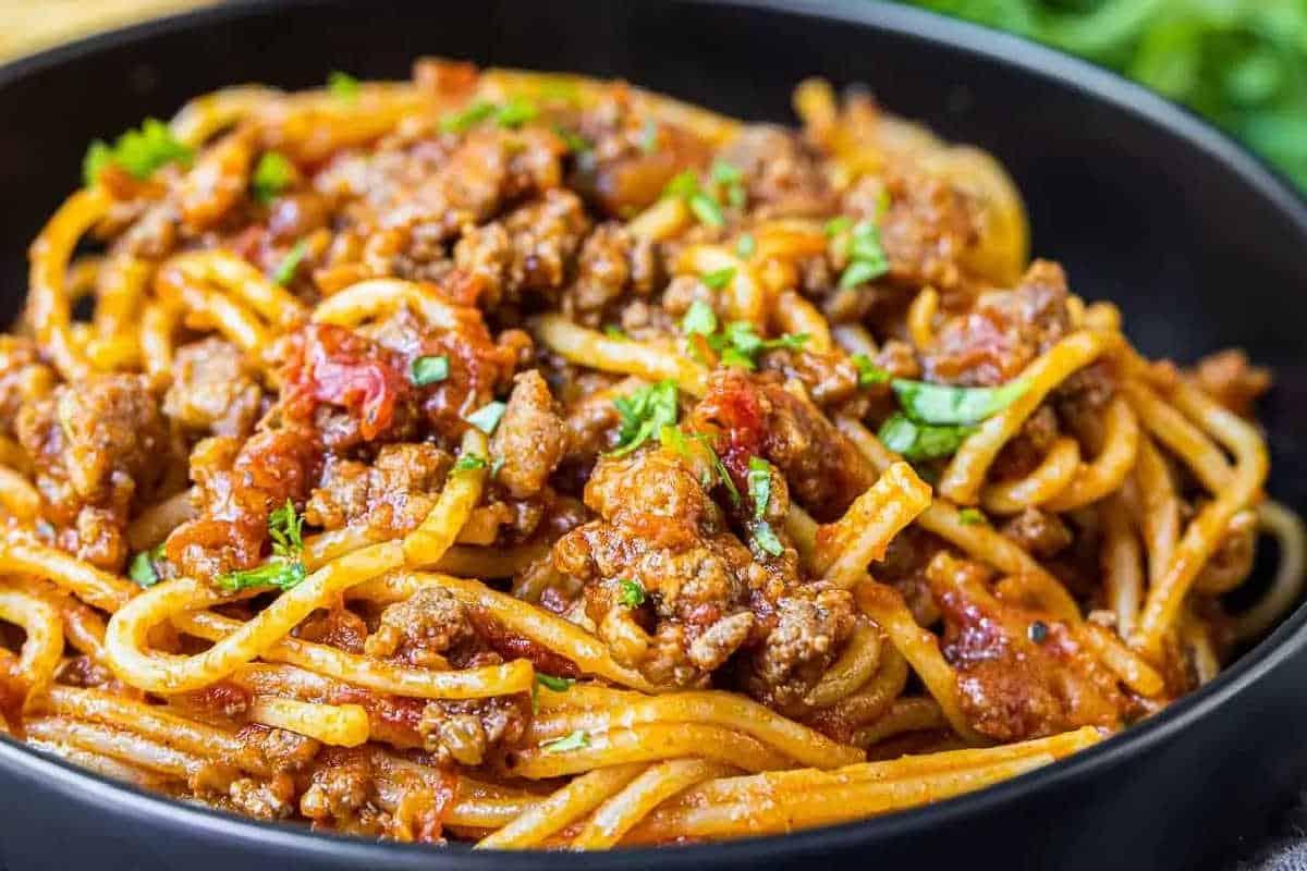 33 einfache und günstige Gerichte (die Ihre Familie auch wirklich essen möchte) 22 Nahaufnahme von Spaghetti, belegt mit Fleischsauce und garniert mit gehackten Kräutern, serviert in einer schwarzen Schale.