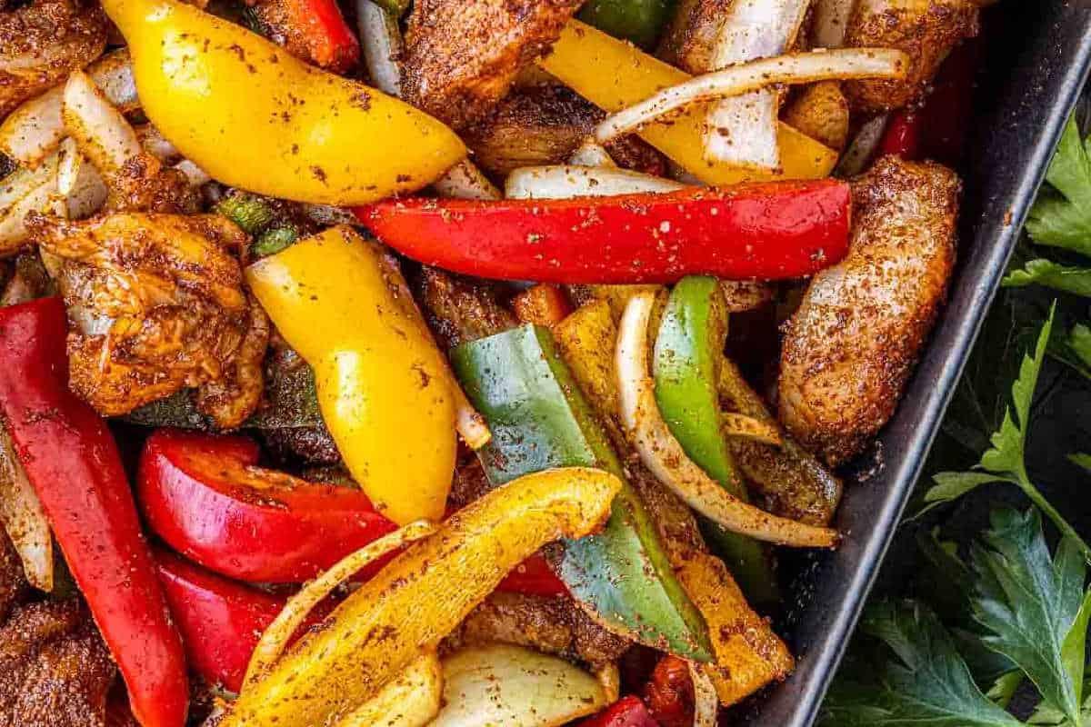 33 einfache und günstige Gerichte (die Ihre Familie auch wirklich essen möchte) 24 Nahaufnahme von gewürzten Hähnchenstreifen, geschnittener roter, gelber und grüner Paprika sowie Zwiebeln auf einem Backblech.