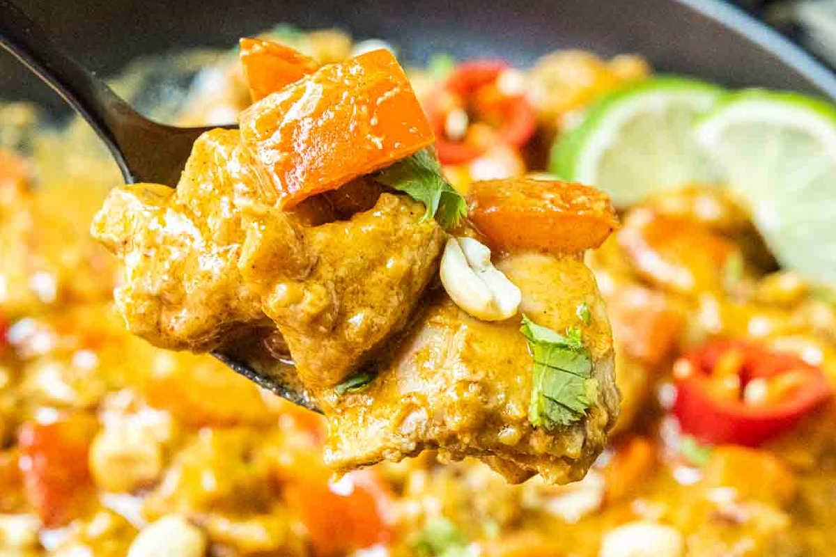 33 einfache und günstige Gerichte (die Ihre Familie auch wirklich essen möchte) 27 Nahaufnahme eines Löffels mit cremigem Erdnuss-Hähnchen-Curry mit Hähnchenstücken, roter Paprika, Erdnüssen, Koriander und einer Soße, mit Limettenscheiben und Chilischoten im Hintergrund.