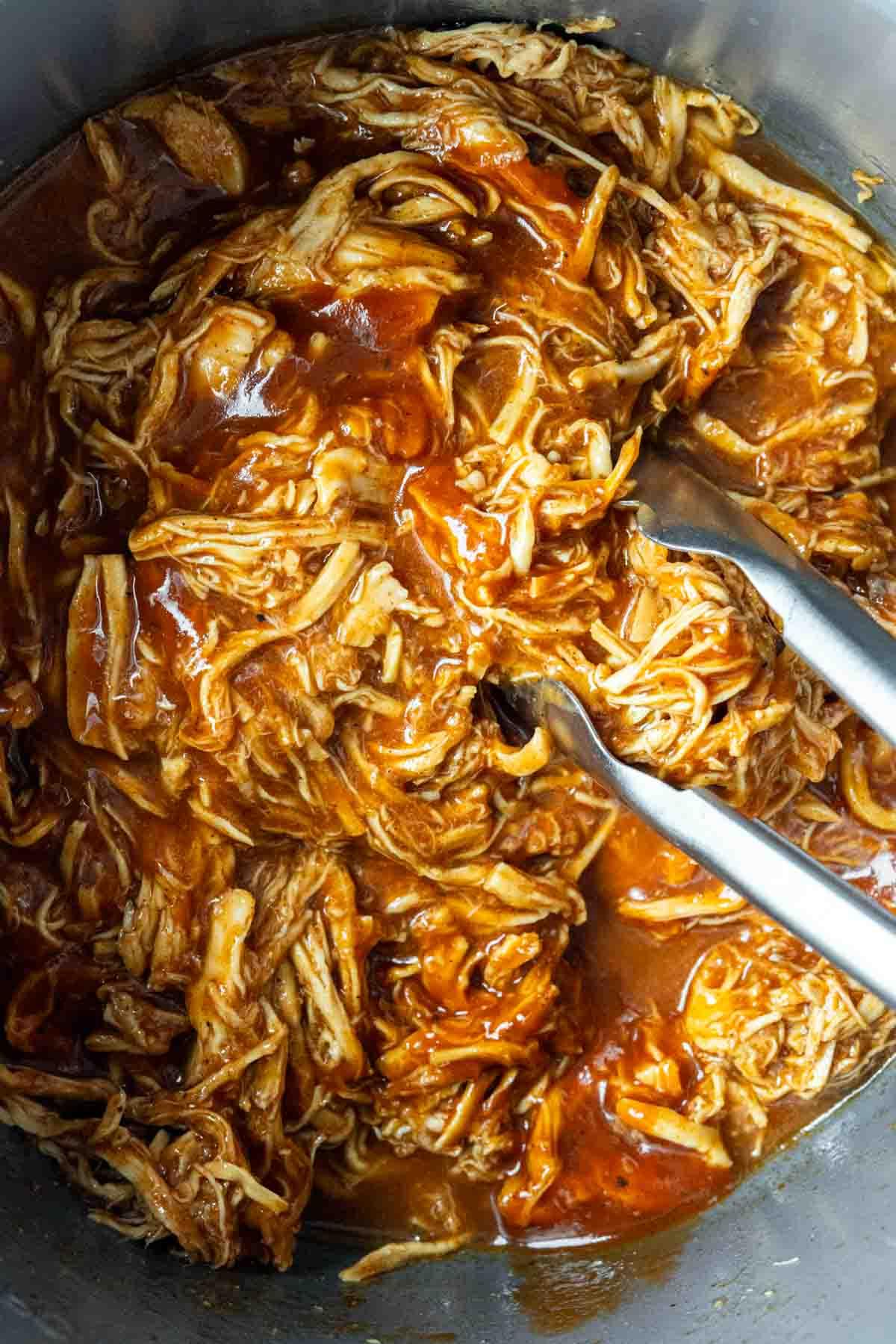 BBQ Pulled Chicken im Slow Cooker 4 Nahaufnahme von gerupftem Barbecue-Hähnchen in einem Metalltopf, das mit Zangen vermischt wird. Das Gericht hat eine satte, rötlich-braune Soße, die das Hähnchen bedeckt.