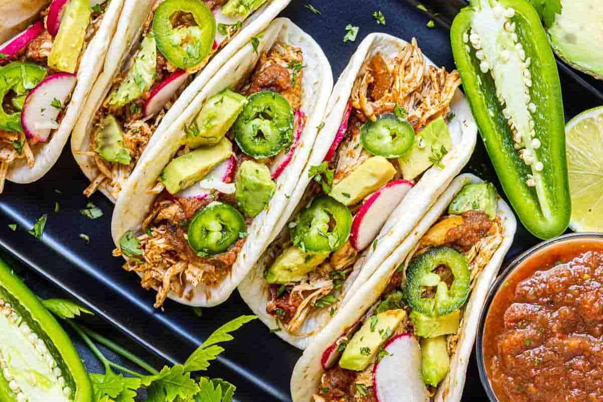 **Schnelle und einfache Hähnchengerichte für vielbeschäftigte Familien** 4 Verschiedene Hühnchen-Tacos mit Avocadoscheiben, Jalapeños und Radieschen auf einem schwarzen Teller, begleitet von Limettenspalten und Salsa.