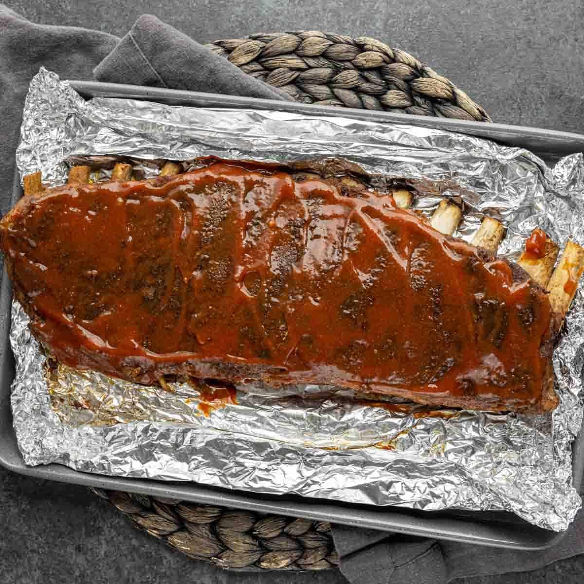 St. Louis Style Spareribs: Langsam gegarte Perfektion 4 Aufsicht auf einen Strang St. Louis-Style Ribs, bedeckt mit Sauce, in Folie gewickelt auf einem Tablett mit Seilgriff.