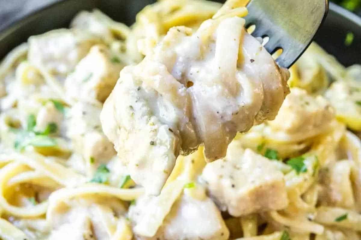 33 einfache und günstige Gerichte (die Ihre Familie auch wirklich essen möchte) 33 Eine Gabel hält einen Bissen cremiger Hähnchen-Alfredo-Pasta, mit Hähnchenstücken, Nudeln und einer weißen Sauce mit Kräutern.
