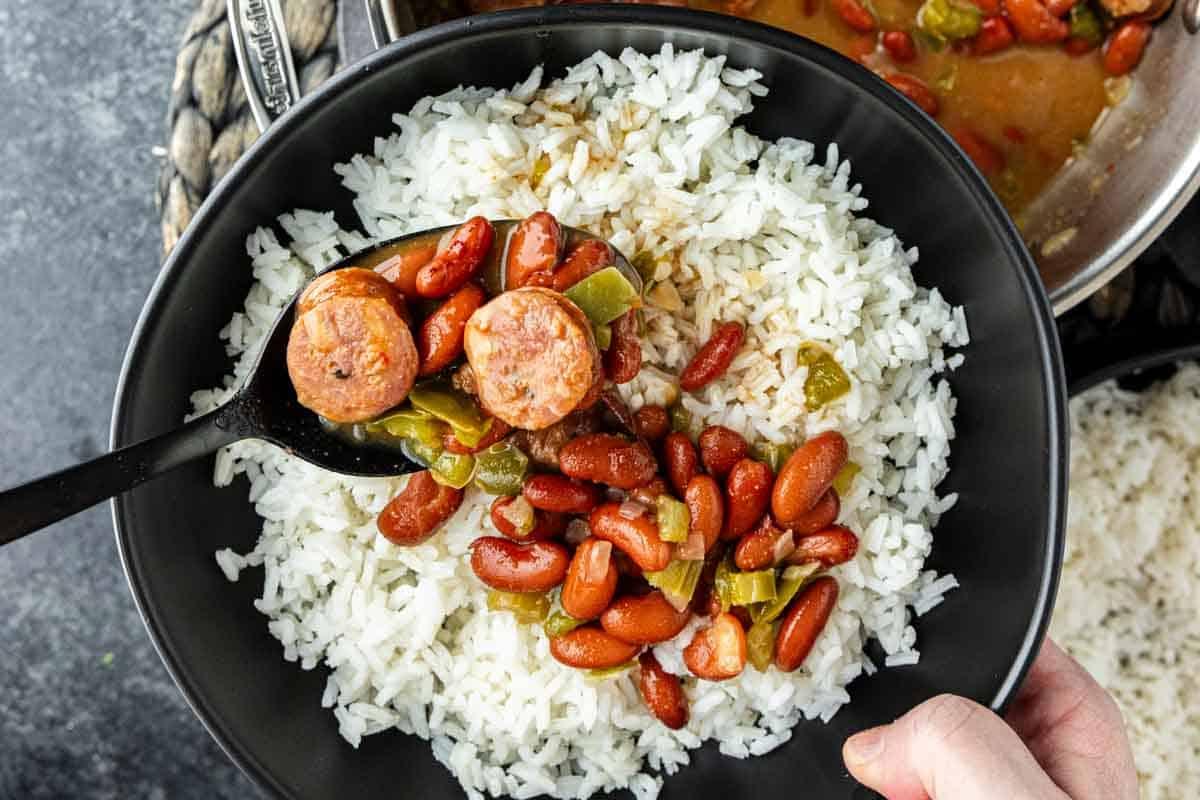33 einfache und günstige Gerichte (die Ihre Familie auch wirklich essen möchte) 34 Eine schwarze Schale gefüllt mit weißem Reis, belegt mit roten Bohnen, Wurstscheiben und grüner Paprika, serviert mit einem Löffel.