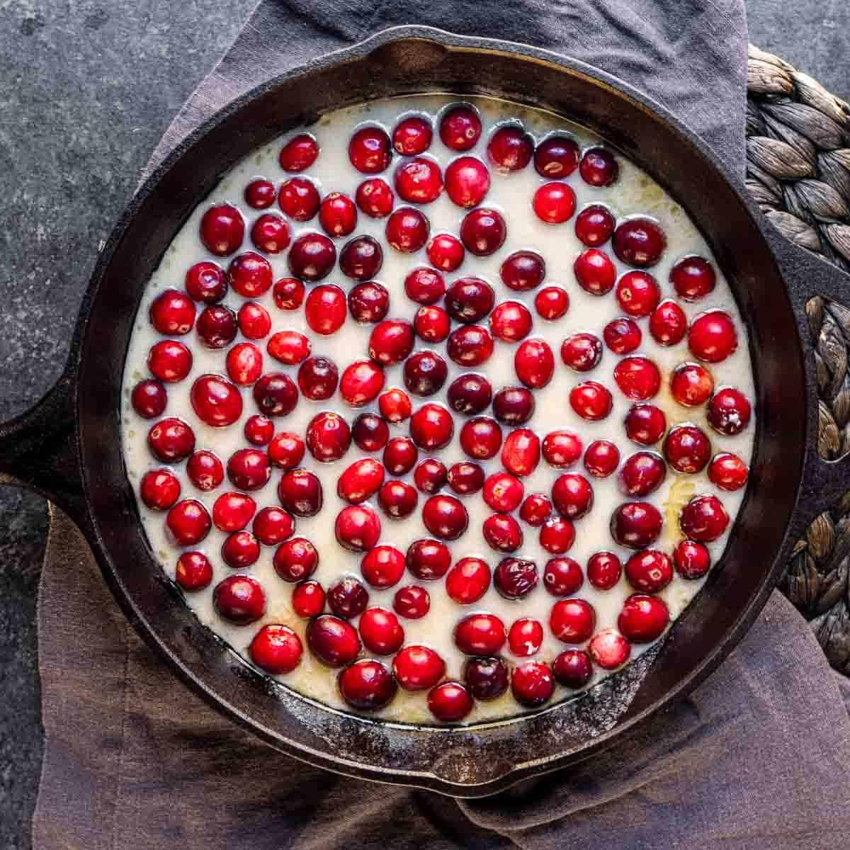 Cranberry-Streuselkuchen 5 Eine Gusseisenpfanne, gefüllt mit Teig, bedeckt mit frischen Preiselbeeren, auf einem dunklen Tuch ruhend.