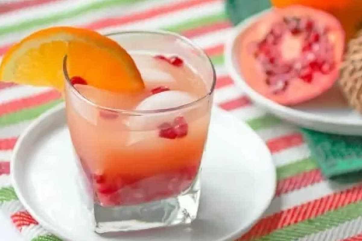 **Ein Galentine’s-Schmaus für deine Mädels** 5 Ein Glas pinkfarbener Saft mit Eiswürfeln, Granatapfelkernen und einer Orangenscheibengarnitur, auf einem weißen Teller auf einem gestreiften Tischtuch. Ein halbierter Granatapfel ist im Hintergrund.