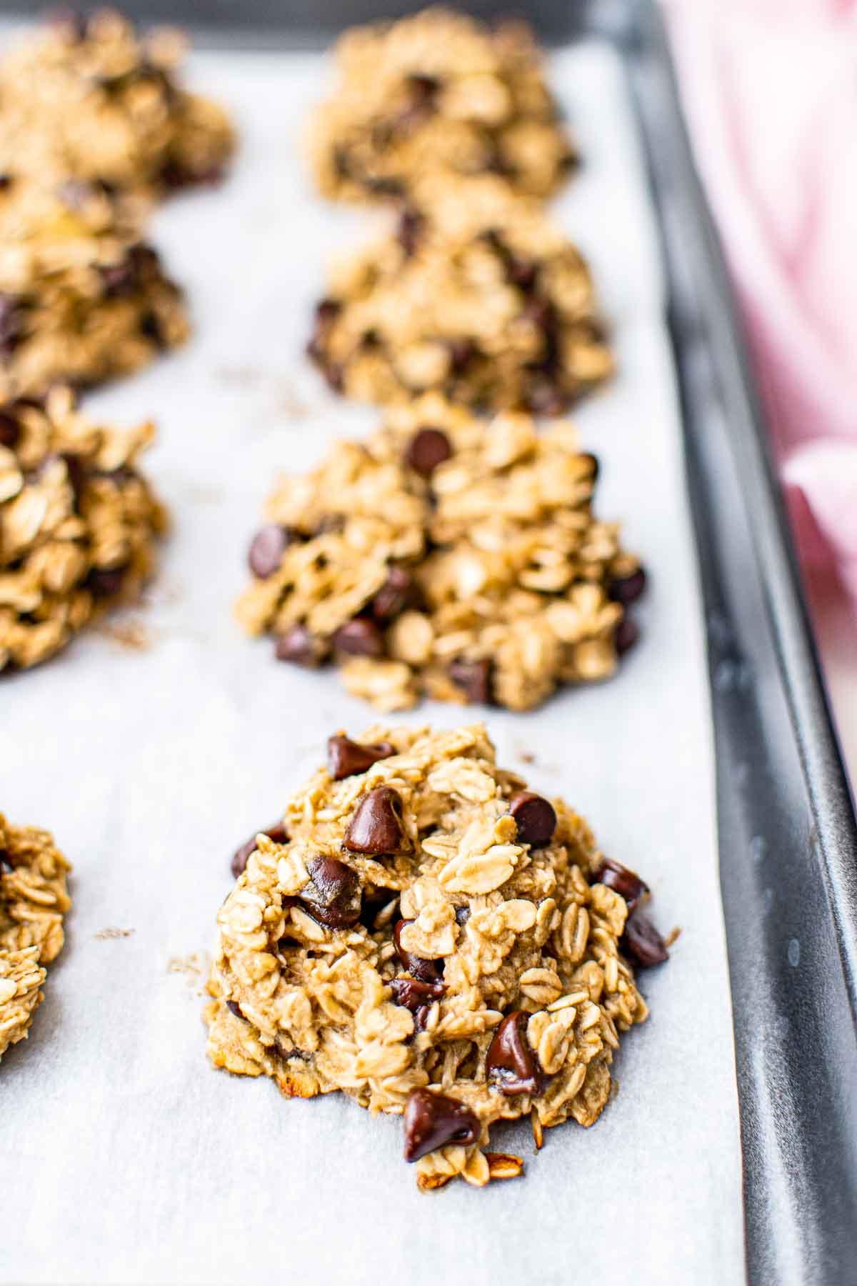 Gesunde Haferflockenkekse – Einfaches Frühstück oder Snack! 5 Ein Backblech mit Backpapier, auf dem mehrere Haferflocken-Schokostückchen-Kekse liegen, mit sichtbaren Haferflocken und Schokostückchen.