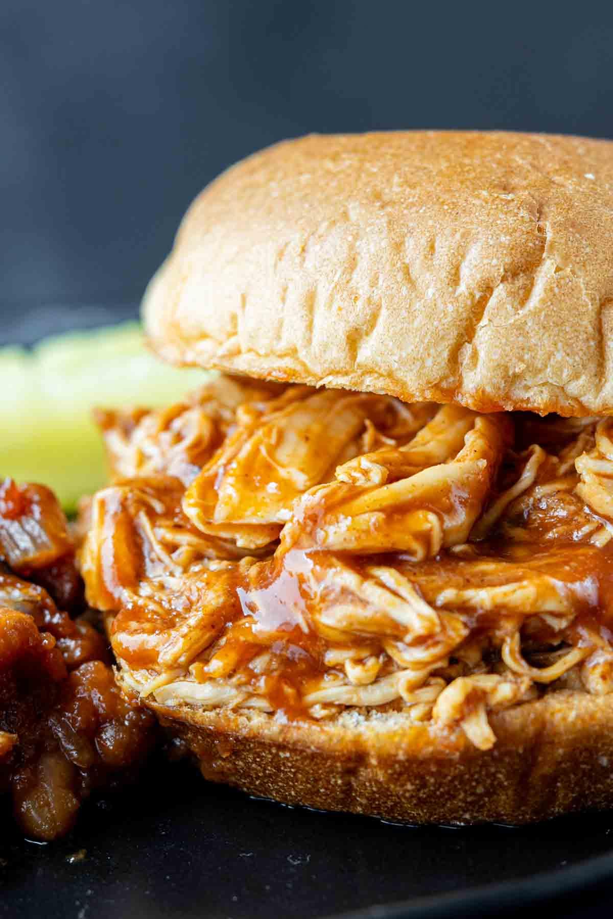 BBQ Pulled Chicken im Slow Cooker 5 Nahaufnahme eines Barbecue-Hähnchen-Sandwiches mit gerupftem Hähnchen in Soße, serviert auf einem Brötchen mit einer Beilage gebackener Bohnen und einer Ananasscheibe.