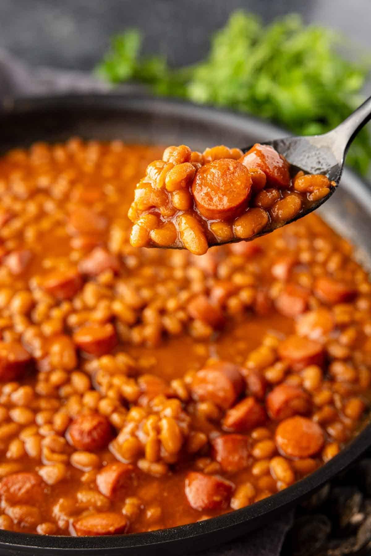 Klassisches Rezept für Würstchen mit Bohnen 6 Ein Löffel hält hausgemachte Würstchen und Bohnen über einer Pfanne mit dem gleichen Gericht.