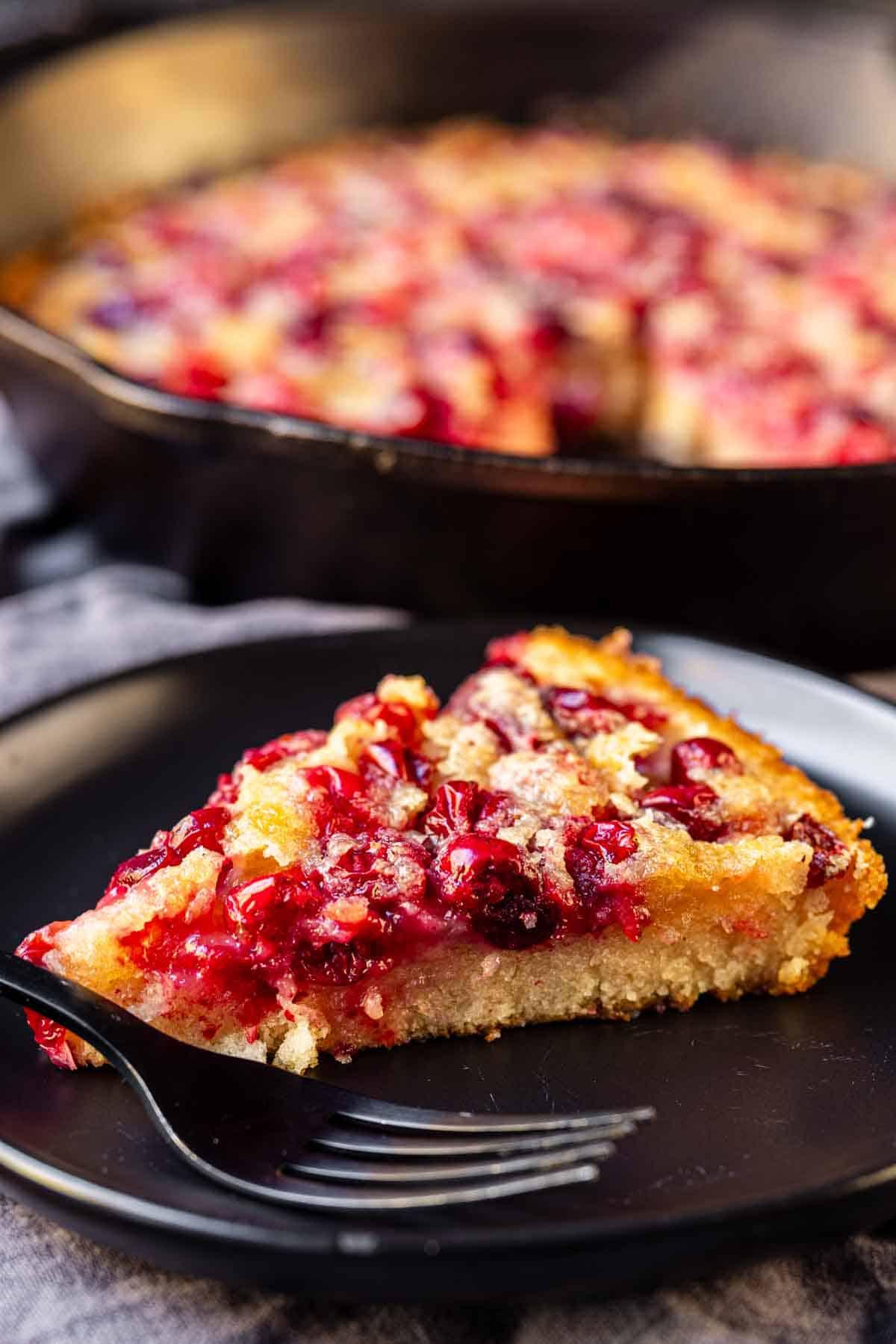 Cranberry-Streuselkuchen 6 Eine Scheibe des beerenbedeckten Desserts wird auf einem schwarzen Teller mit einer Gabel serviert, vor dem restlichen Dessert in einer Pfanne.
