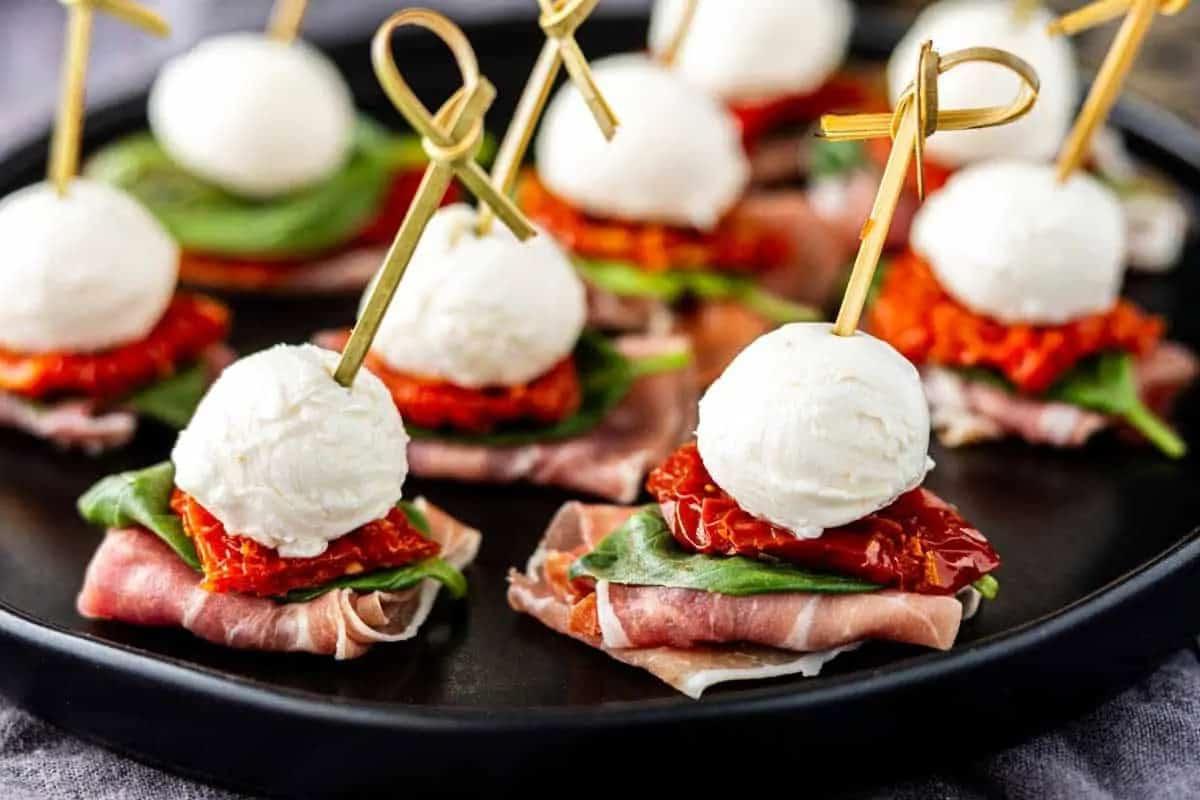 **Ein Galentine’s-Schmaus für deine Mädels** 6 Vorspeisen auf Spießen mit Mozzarella-Kugeln, Basilikum, getrockneten Tomaten und Prosciutto auf einem schwarzen Tablett.