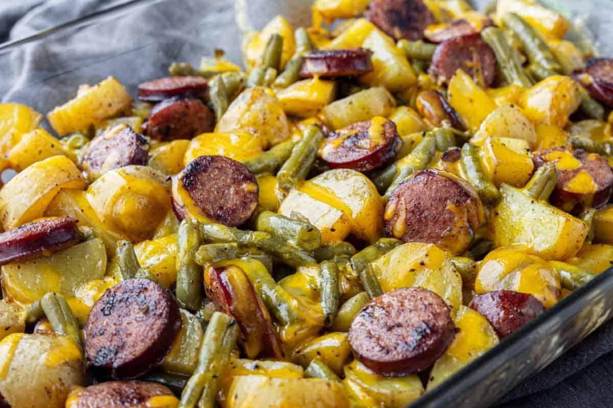 33 einfache und günstige Gerichte (die Ihre Familie auch wirklich essen möchte) 6 Ein gebackenes Gericht mit geschnittenen Würstchen, gelben Kartoffeln, grünen Bohnen und geschmolzenem Käse in einer Glasbackform.