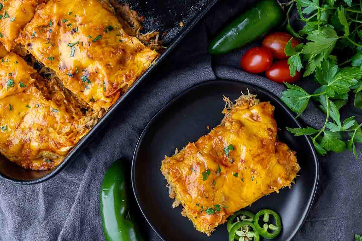 Festliche Abendessenrezepte für den Cinco de Mayo 6 Ein Teller Enchiladas mit Käse und Garnitur, daneben frische Jalapeños, Tomaten und Kräuter, serviert auf einem dunklen, strukturierten Hintergrund.