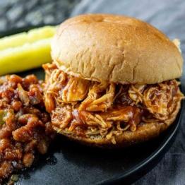 BBQ Pulled Chicken im Slow Cooker 6 Ein Pulled Chicken Sandwich mit Barbecue-Soße liegt auf einem schwarzen Teller, begleitet von gebackenen Bohnen und einigen Gewürzgurkenstücken.