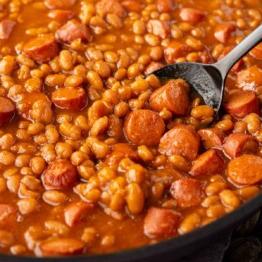 Klassisches Rezept für Würstchen mit Bohnen 7 Nahaufnahme von gebackenen Bohnen, gemischt mit geschnittenen Würstchen in einer Pfanne, mit einem Löffel, der etwas herausnimmt.