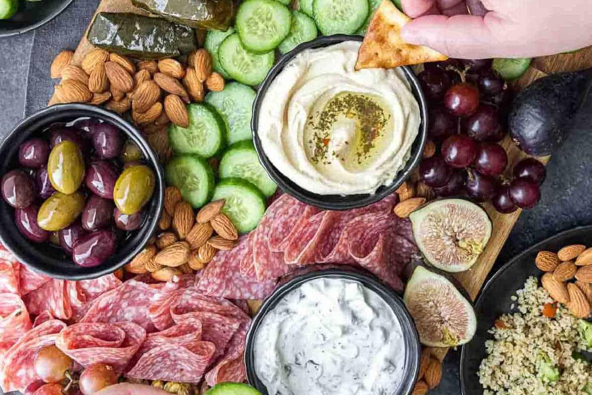 **Ein Galentine’s-Schmaus für deine Mädels** 7 Eine Hand taucht einen Pitachip in Hummus auf einer Platte mit geschnittenen Gurken, Oliven, Mandeln, Feigen, Trauben, Wurstwaren und einer Schüssel Tzatziki.