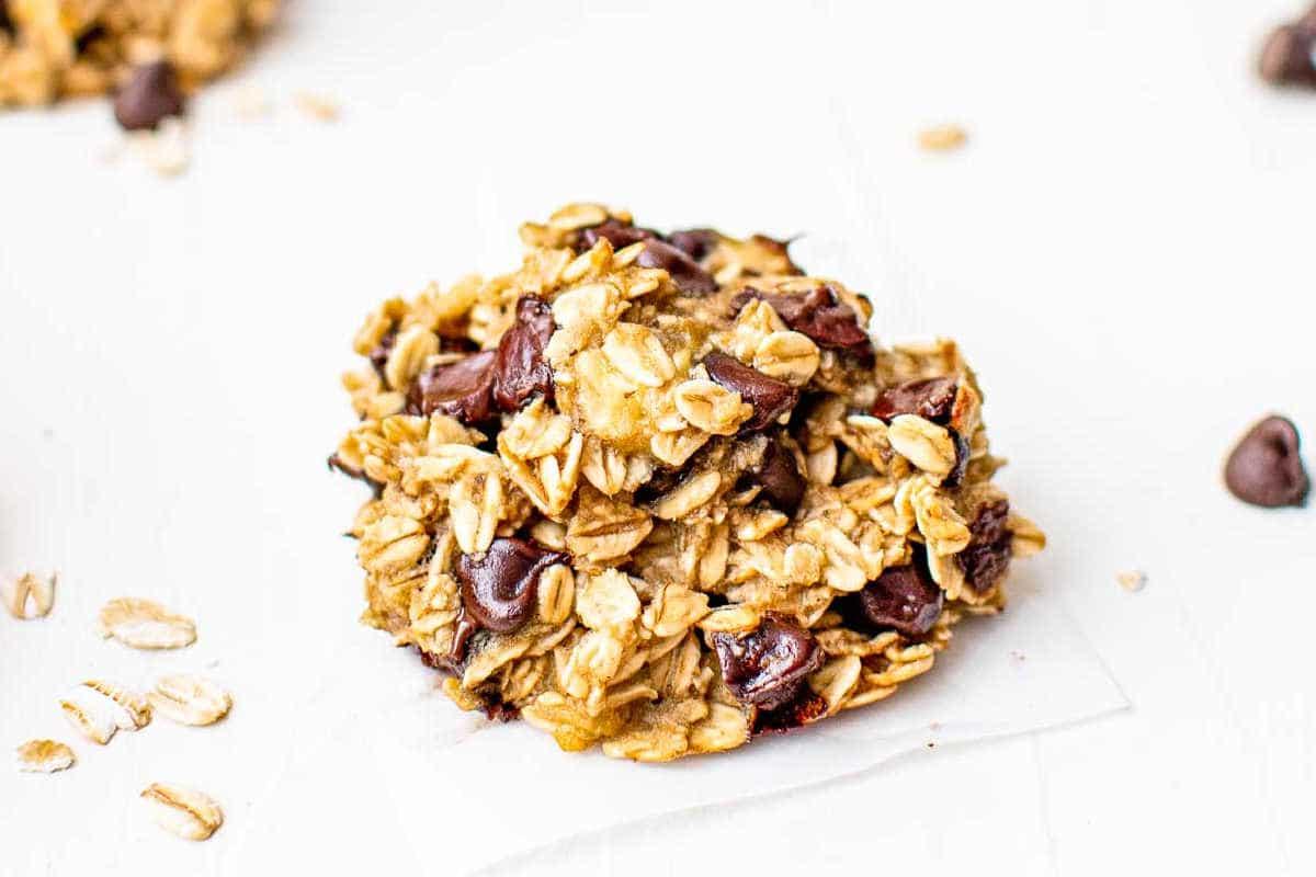Gesunde Haferflockenkekse – Einfaches Frühstück oder Snack! 7 Nahaufnahme eines Haferflockenkekses mit Schokostückchen auf einer weißen Oberfläche, umgeben von verstreuten Haferflocken und Schokostückchen.