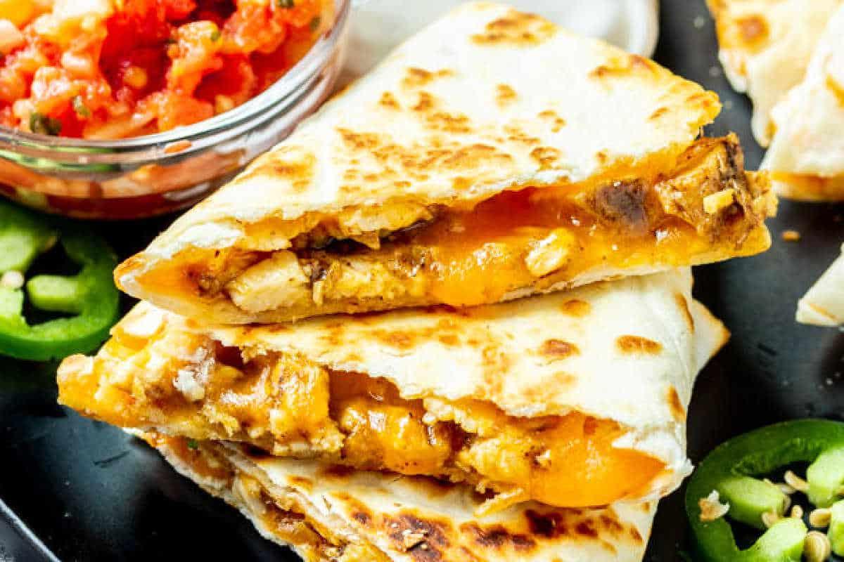 Festliche Abendessenrezepte für den Cinco de Mayo 7 Nahaufnahme einer Hähnchen-Quesadilla mit geschmolzenem Käse, serviert mit Salsa auf einem schwarzen Teller, garniert mit geschnittenen Jalapeños.