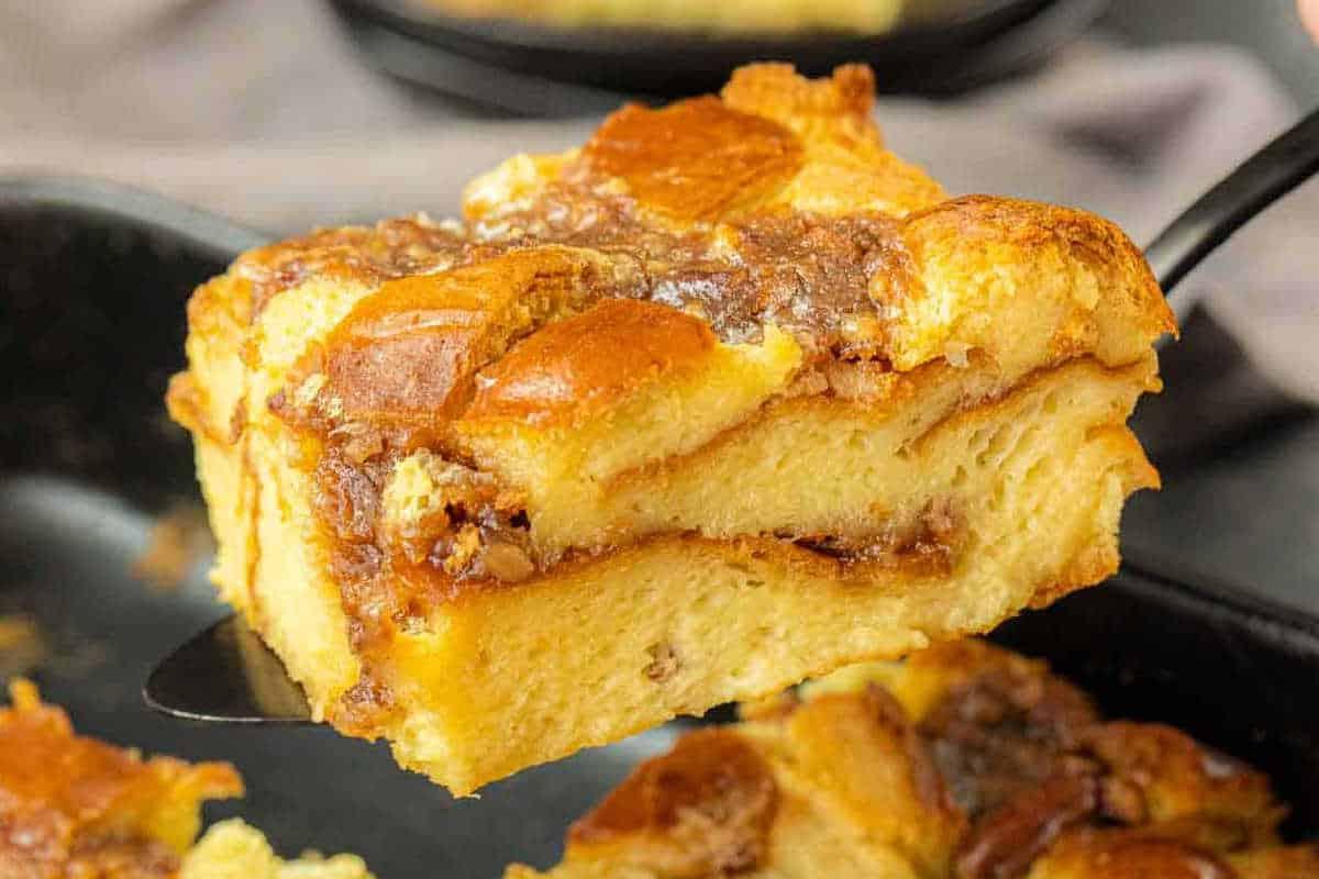 Unwiderstehliche Desserts mit Pekannusskuchenfüllung 7 Nahaufnahme einer Scheibe Brotpudding mit goldbrauner Kruste und Zimtwirbeln, die mit einem Spatel aus einer Pfanne gehoben wird.