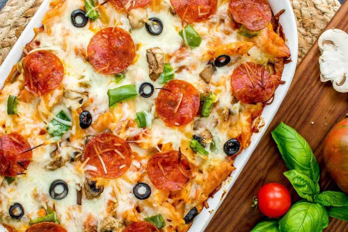 33 einfache und günstige Gerichte (die Ihre Familie auch wirklich essen möchte) 8 Eine frisch gebackene Pizza, belegt mit Peperoni, grüner Paprika, Oliven, Pilzen und geschmolzenem Käse, steht auf einem Holzbrett neben Basilikumblättern und Kirschtomaten.