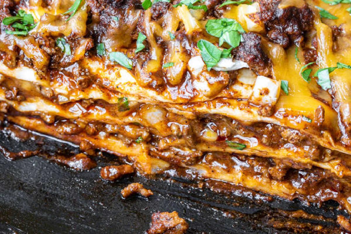 Festliche Abendessenrezepte für den Cinco de Mayo 8 Nahaufnahme eines geschichteten Lasagne-ähnlichen Gerichts mit geschmolzenem Käse und Fleischsauce, garniert mit gehackten Kräutern.