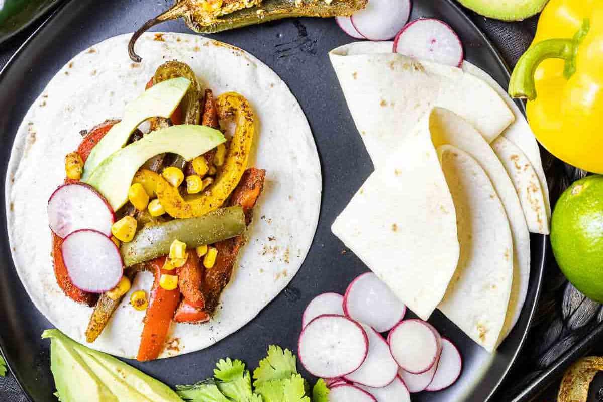 Festliche Abendessenrezepte für den Cinco de Mayo 10 Festliche offene Tortilla mit sautiertem Gemüse, Avocadoscheiben und Radieschen, begleitet von Limettenspalten und Koriander auf einem schwarzen Teller.